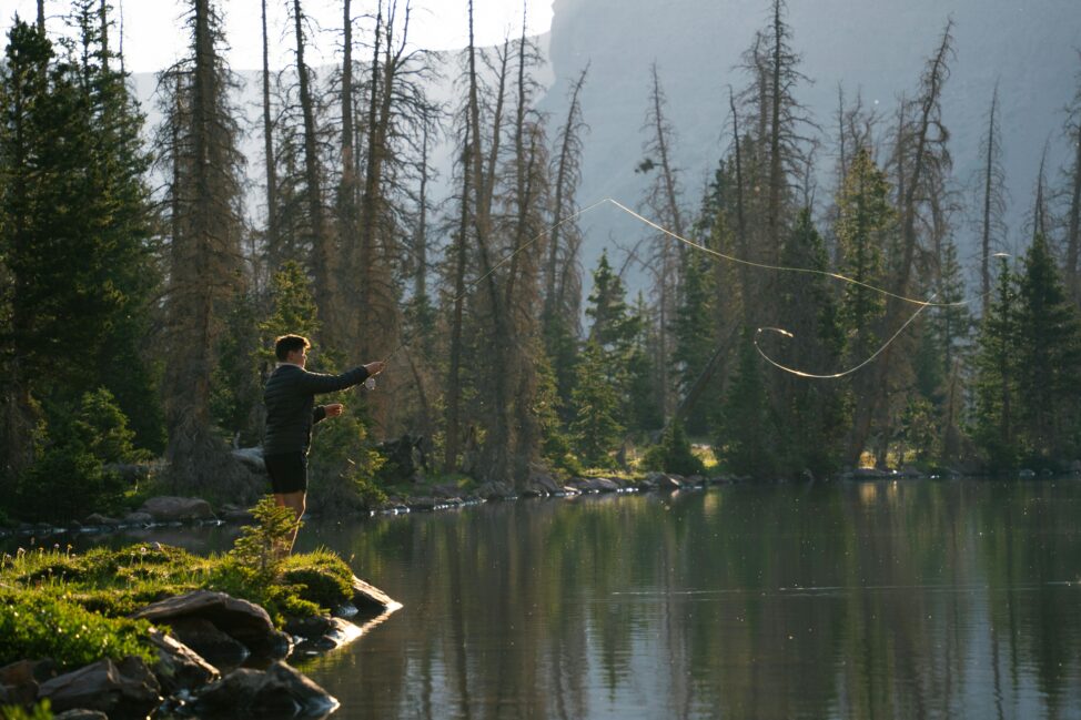 a man standing on green grass casting a fishing line into water. The man and water are in the woods with mountains in the background. | Finding Purpose Beyond Medicine: What Really Matters?